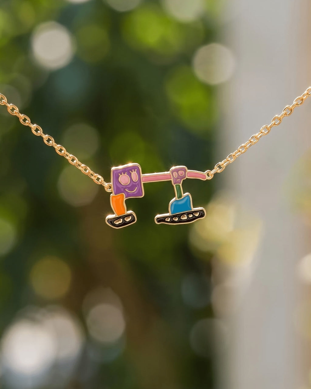 Two block-shaped enamel drawing charms on a gold-tone chain against a green background.