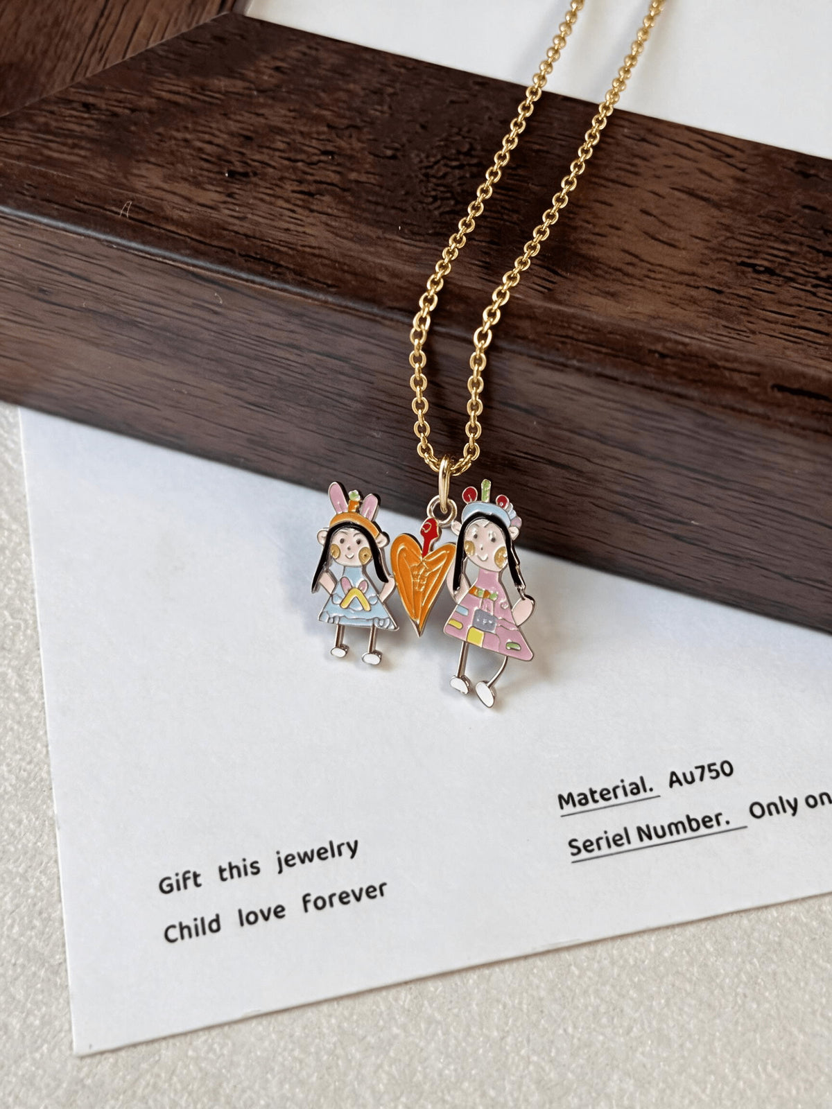 Two-person enamel drawing pendant with a heart between the figures, hanging beside a wooden frame.