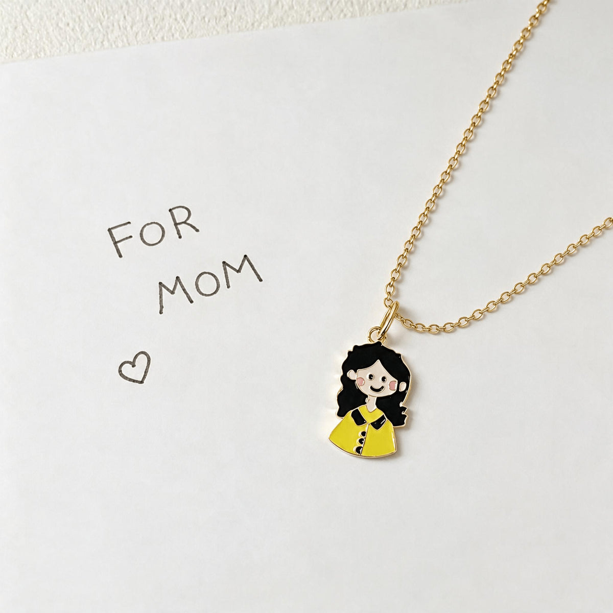 Small enamel girl portrait pendant on a gold-tone chain beside a handwritten For Mom note.